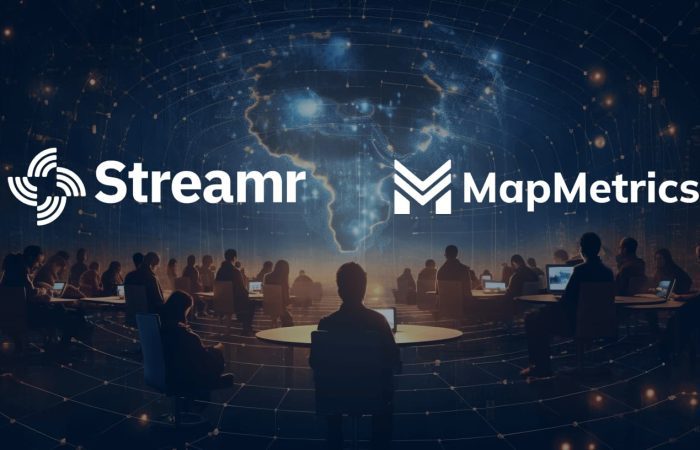 People in a dimly lit room using computers, with a 3D holographic world map showcasing p2p data sharing connections. Streamr logo on the top-left and MapMetrics logo on the top-right, symbolizing their collaboration.