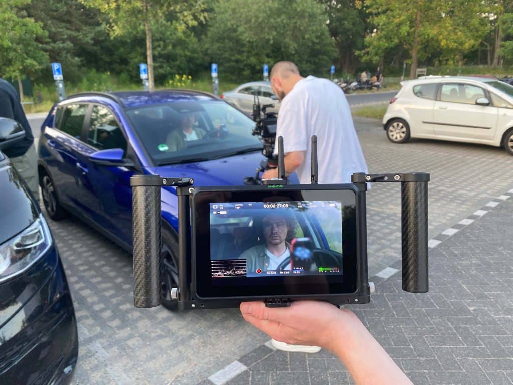 **Alt Text:**A person holding a camera monitor displaying a man in a car, with a film crew working around a blue car in the background. Other parked cars and trees are visible in the parking lot setting.